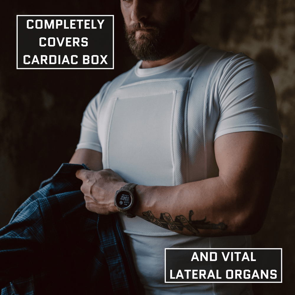 A man wears the Premier Body Armor Everyday Armor T-Shirt 360 (Armored), pulling down his plaid shirt to reveal a white vest labeled "Completely covers cardiac box and vital lateral organs.