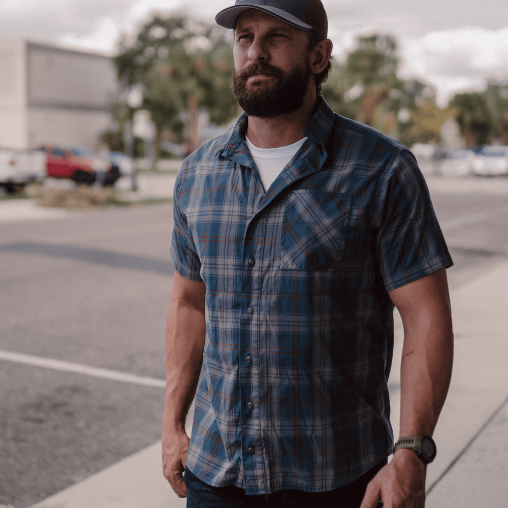 A bearded man in a Premier Body Armor Everyday Armor T-Shirt 360 under a blue plaid short-sleeve shirt, dark jeans, black cap, and wristwatch walks on a sidewalk beside a street lined with cars and trees.