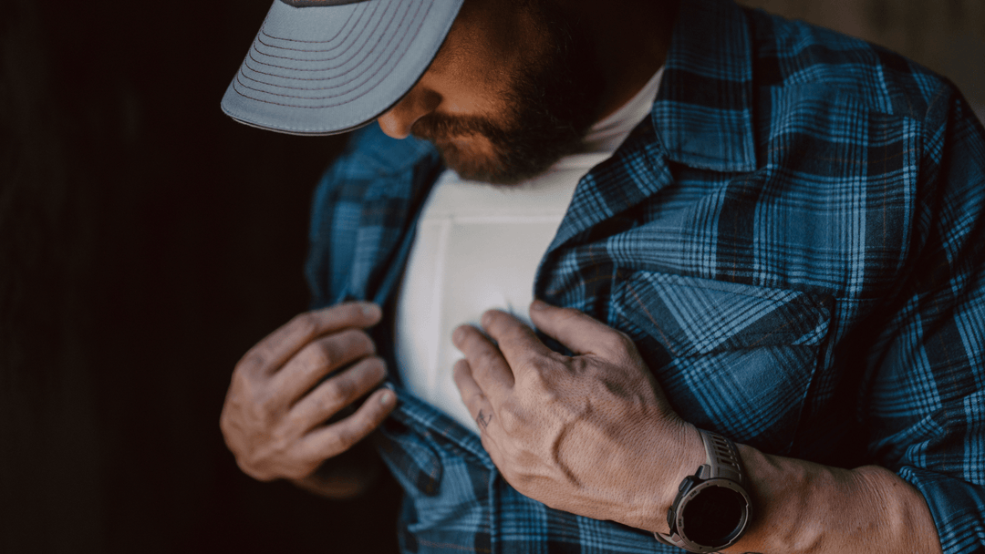 A man wearing a blue plaid shirt and a gray baseball cap looks down while holding the front opening of his shirt with both hands. He has a smartwatch on his left wrist and is wearing a white t-shirt underneath.