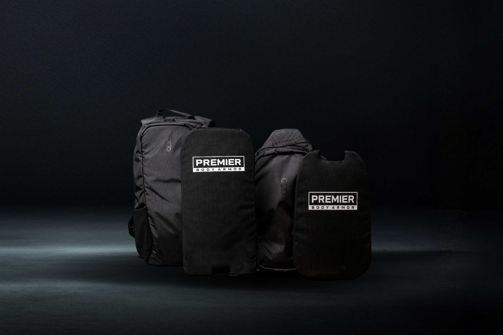 Four black Premier Body Armor products are displayed against a dark background. From left to right, there are a backpack, two armor panels, and a vest. The products have visible Premier Body Armor branding.