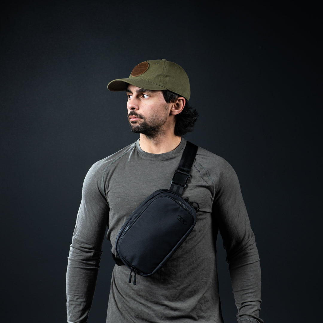 A bearded man wearing a green cap and gray long-sleeve shirt looks left. His black crossbody bag, potentially made of ballistic material, complements the dark background. Featured inside is the Eberlestock Fade Flex Level IIIA Insert by Premier Body Armor.