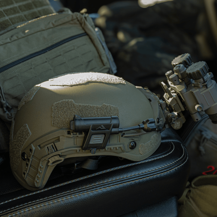 The Fortis Ballistic Helmet by Premier Body Armor, featuring night vision goggles and constructed with lightweight ballistic fibers, rests on a vehicle seat. Its olive green design includes various attachments, while military gear like a backpack in the background enhances the tactical scene.