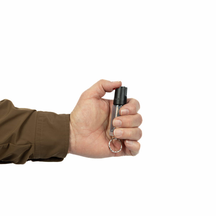 A person in a brown long-sleeve jacket holds a Fox Labs FoxFire Inferno Twin Pack black keychain pepper spray with a keyring, set against a plain white background.