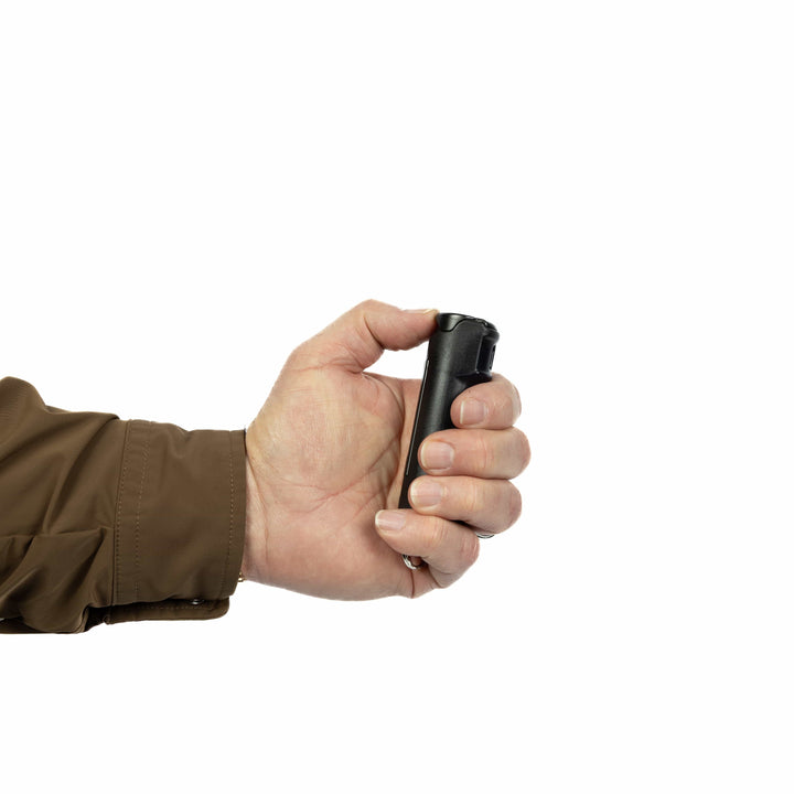 A person in a brown jacket holds a Fox Labs Personal Pocket Unit keychain pepper spray canister by Fox Labs in their right hand against a plain white background.