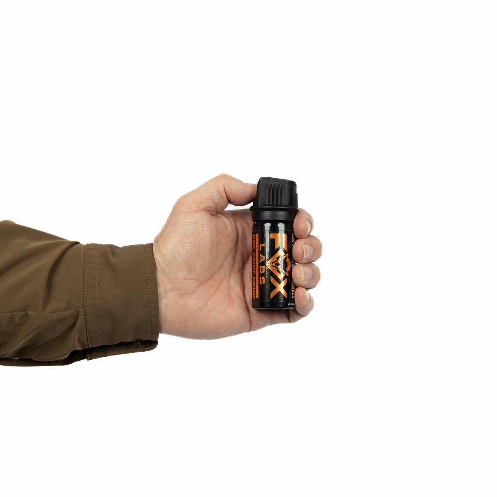 A person in a brown long-sleeve shirt holds a Fox Labs One Point Four 1.5oz Pepper Spray canister with orange and white text against a plain white background.