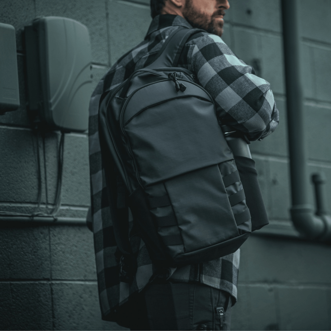 A person wearing a black and gray checkered jacket stands against a gray brick wall, carrying the sleek Vertx Urban Ghost Pack. The image highlights the backpack's 420D ripstop fabric and innovative concealed carry compartment, focusing on the person's upper body.