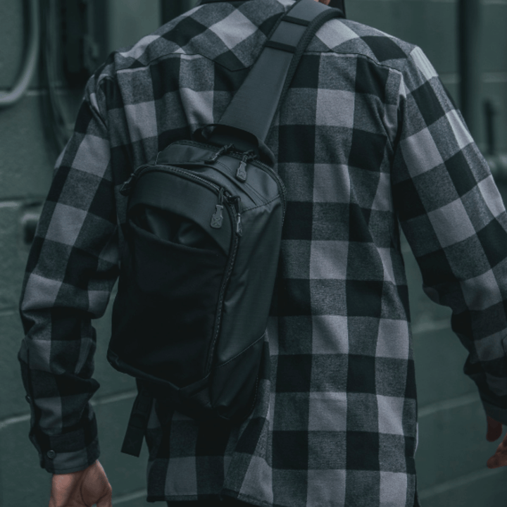 A person wearing a black and white plaid shirt demonstrates the stealth functionality of their Vertx Urban Ghost Sling. The dark wall with hardware fixtures enhances the depth, highlighting the everyday carry essentials of this sleek crossbody bag and the timeless shirt pattern from behind.