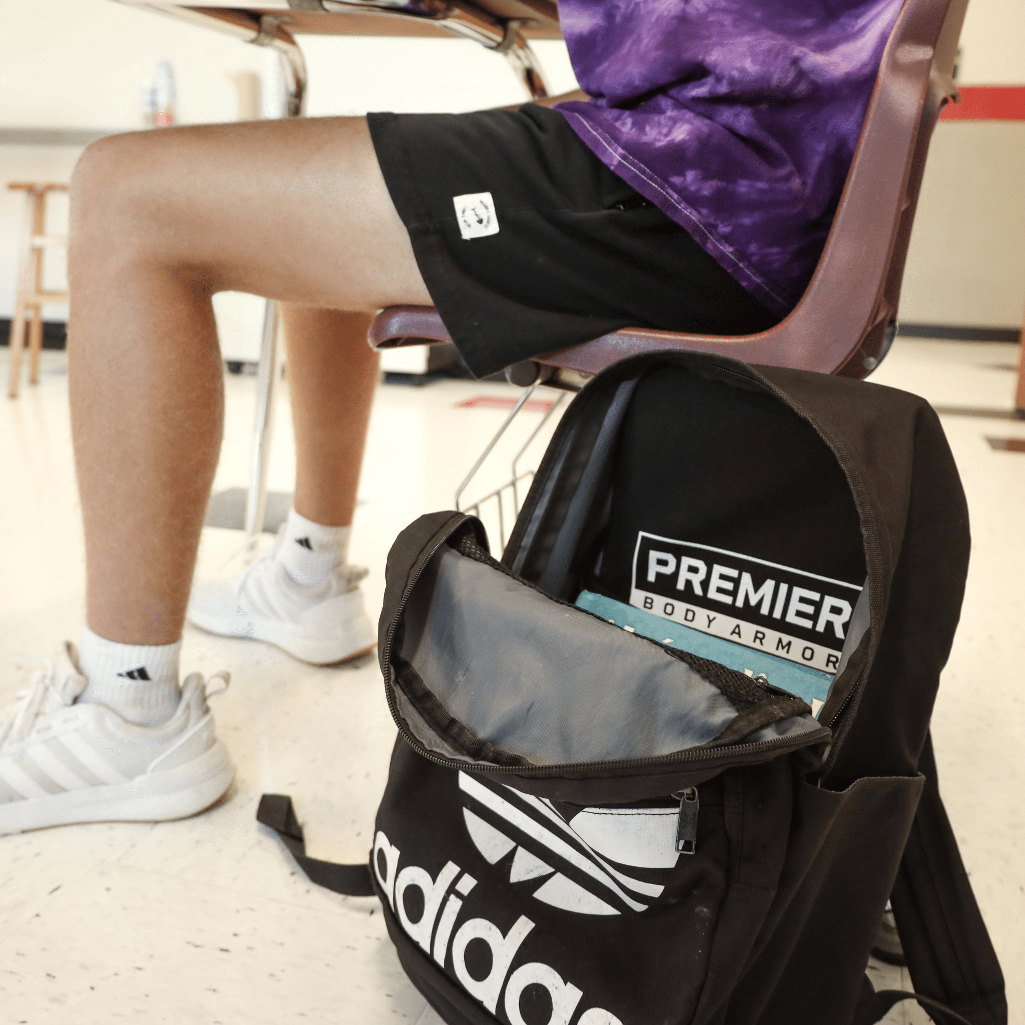 A person wearing a purple shirt and black shorts sits in a chair with crossed legs. A black Adidas backpack with visible "Premier Body Armor" is on the floor, partially open, revealing a blue interior. The setting appears to be a classroom.