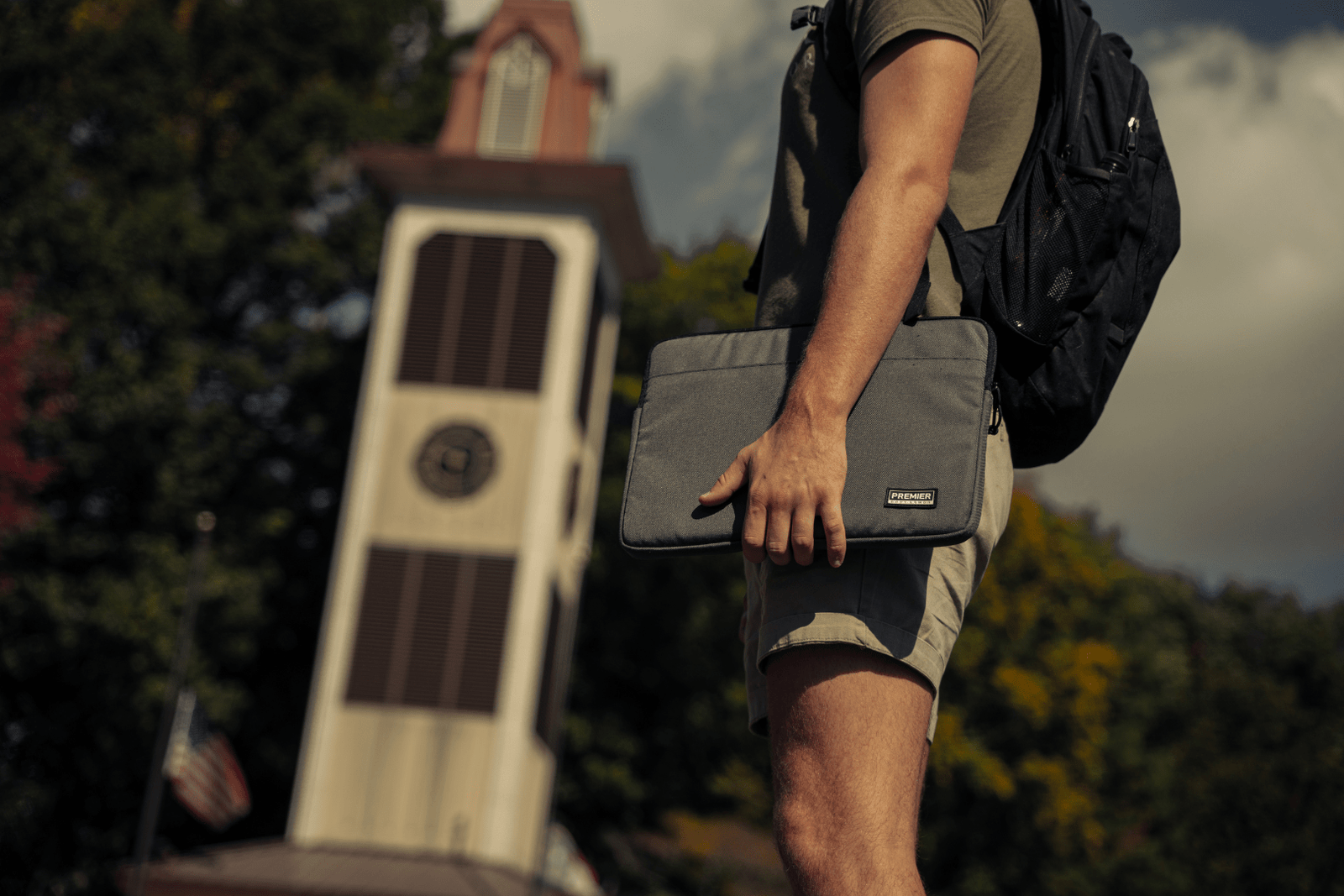 A person holding a grey laptop sleeve stands outdoors in front of a tower clock on a sunny day. The person is wearing a green shirt, beige shorts, and a black backpack. Trees and partly cloudy skies are visible in the background.