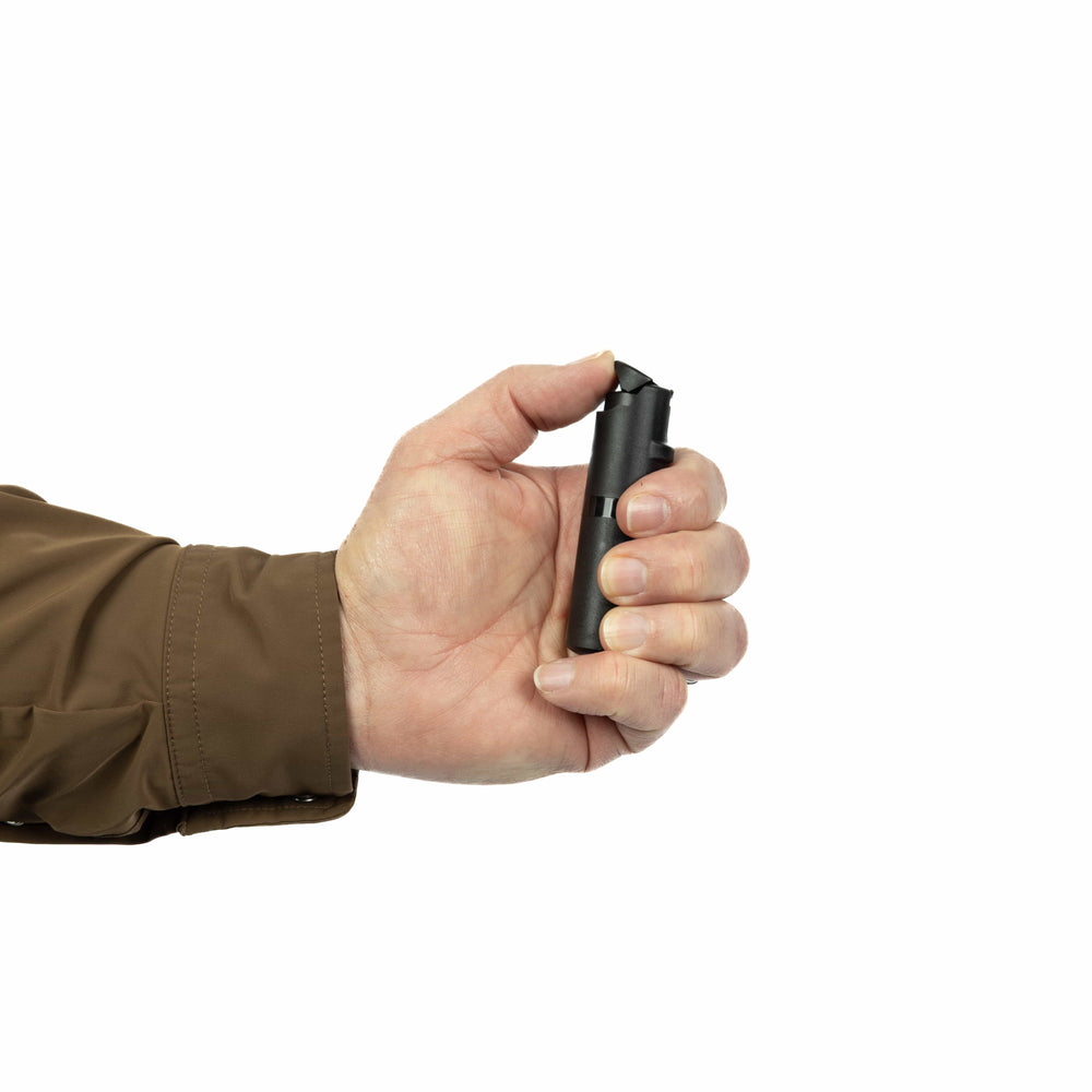 A person in a brown jacket holds a POM Clip Pepper Spray by POM in their right hand against a white background, highlighting this compact self-defense tool for everyday carry.