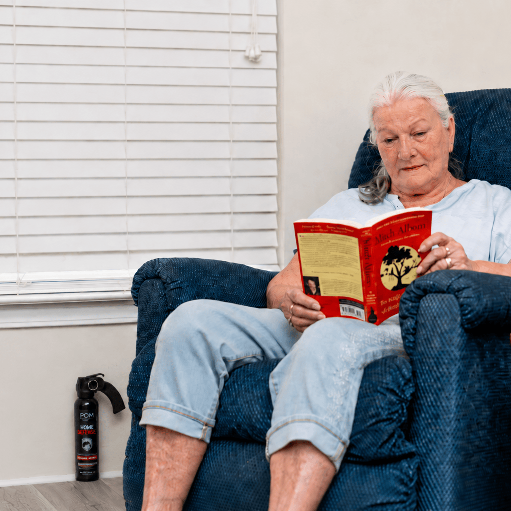 An older woman with long gray hair, wearing a light blue shirt and rolled-up jeans, reads a red book in a blue recliner. Beside her on the floor is POM Home Defense Pepper Spray by POM, near a window with closed white blinds.