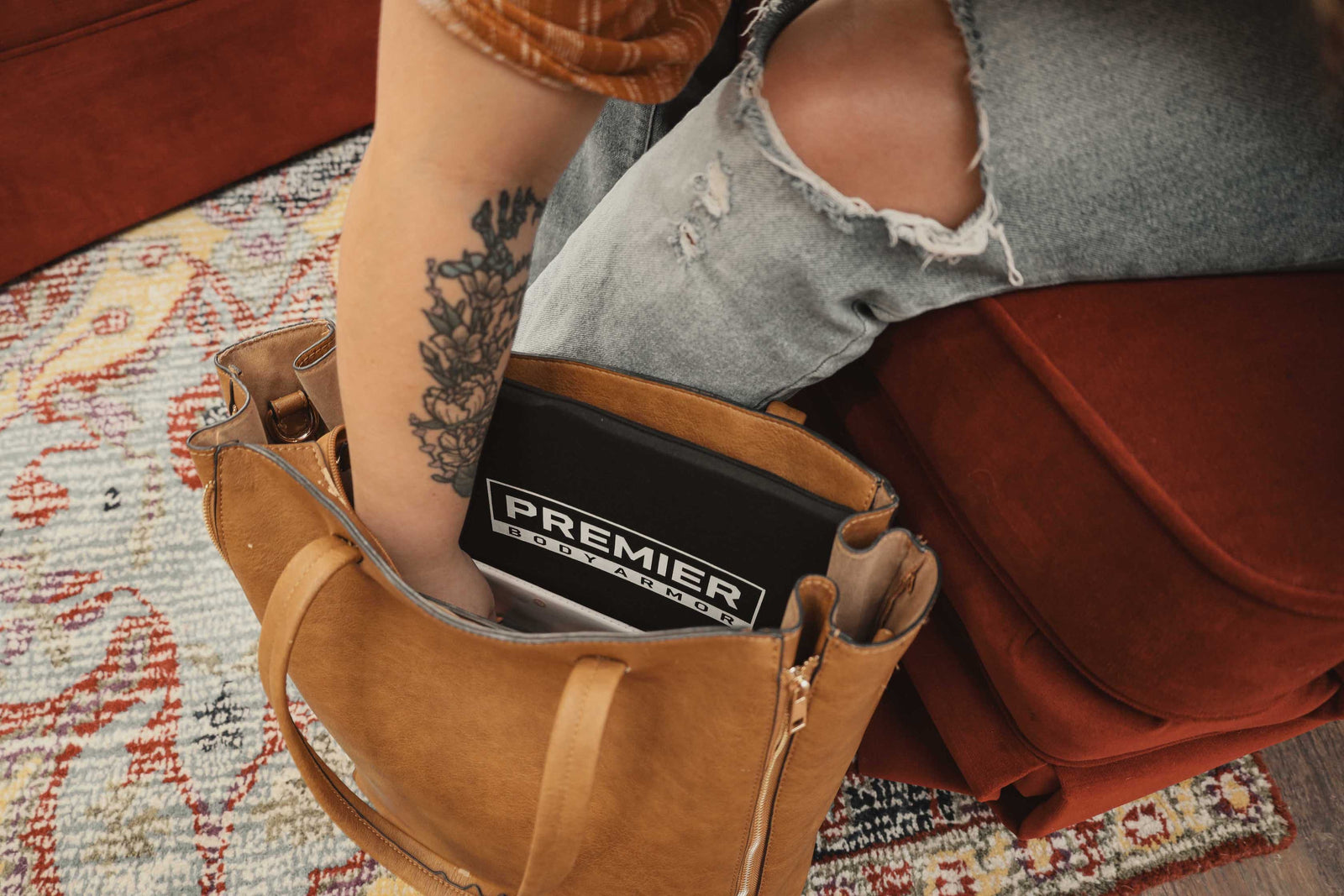 A person with a floral tattoo reaches into a tan bag on a red cushioned seat, retrieving an item labeled "PREMIER." The person is wearing a brown shirt and ripped jeans. The background includes part of a patterned rug.