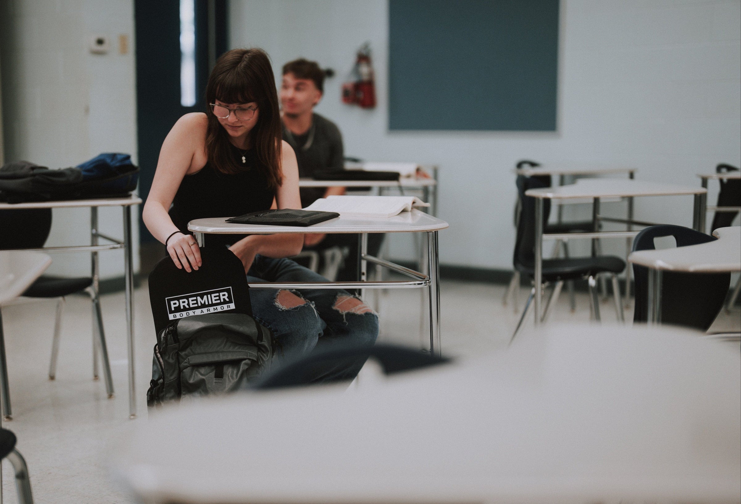 Person sitting at a desk in a classroom with a backpack and Premier Body Armor bulletproof backpack insert on the floor.