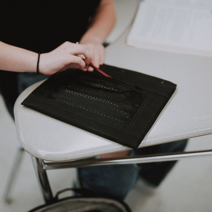 In a light-colored classroom, a person wearing a black shirt opens the Premier Body Armor Level IIIA 3-Ring Pencil Pouch. 