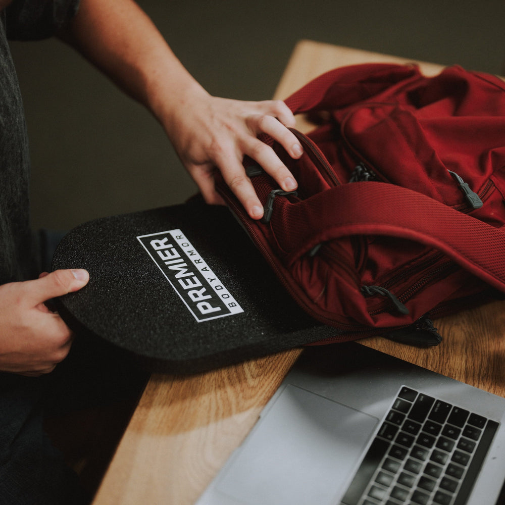 A person puts a black skateboard and a Premier Body Armor 10x15 Special Threat Rifle-Rated Backpack Insert into a red backpack on a wooden table. An open laptop and another person reading a book are also on the table.