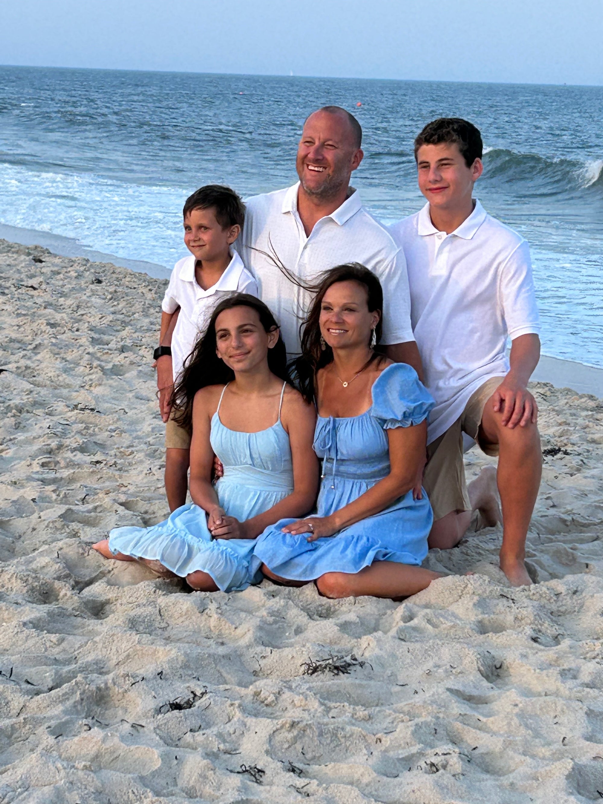 A family of five is posed for a photo on a sandy beach near the ocean. They are all dressed in white and light blue clothing. Two adults are seated in front with two children, while one adult and an older child are standing behind them. The weather appears clear.