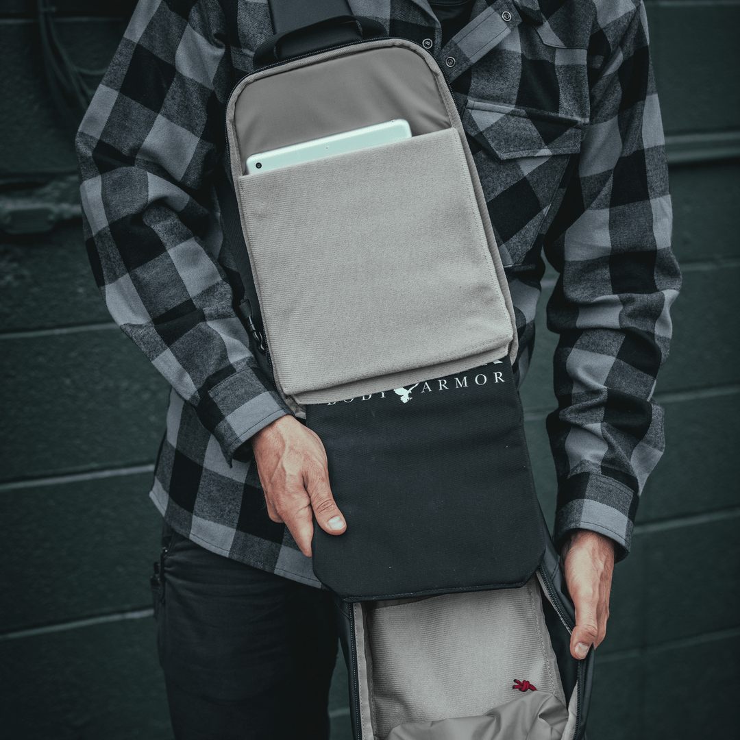 A person wearing a black and gray checkered shirt is holding the Armored Vertx Ghost Sling Bundle by Vertx/Premier, showcasing its tactical utility with a tablet visible in an exterior pocket. The image captures the lower torso and hands, exemplifying both style and function.