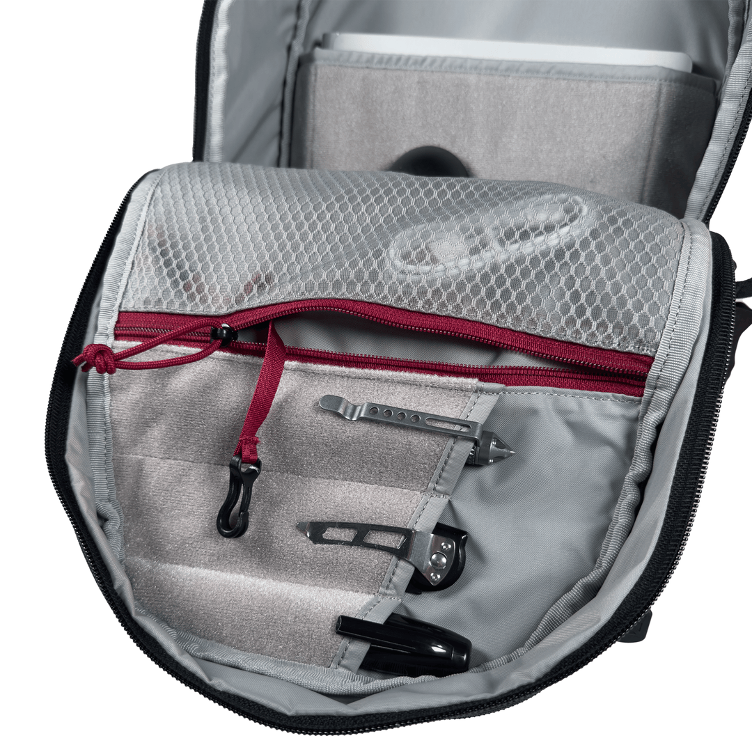 Open the Vertx Ready Pack X backpack by Vertx, revealing a gray interior with a mesh zippered pocket, red zipper pull, and everyday carry items such as paper clips and a black pen neatly organized in loops and pockets.