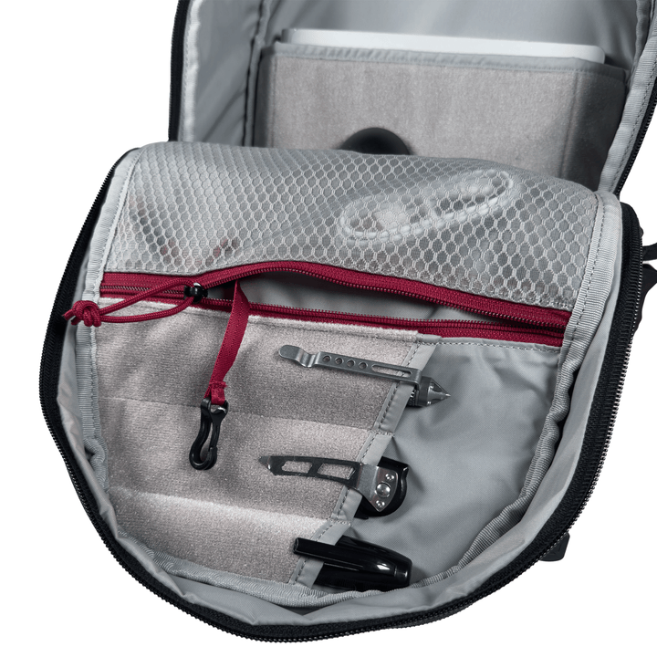Open the Vertx Ready Pack X backpack by Vertx, revealing a gray interior with a mesh zippered pocket, red zipper pull, and everyday carry items such as paper clips and a black pen neatly organized in loops and pockets.