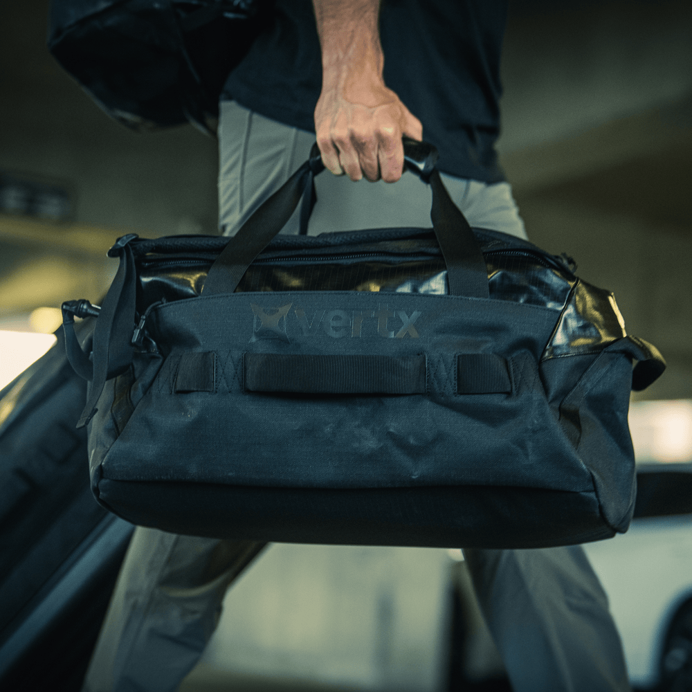 In a parking garage, someone can be seen carrying a Vertx RLT Duffel. The durable black bag, designed with weather-resistant materials, prominently features the "Vertx" logo and provides customizable storage solutions. As they move past the parked cars in the background, the individual is wearing gray pants and easily carries the duffel using its handles or shoulder strap.