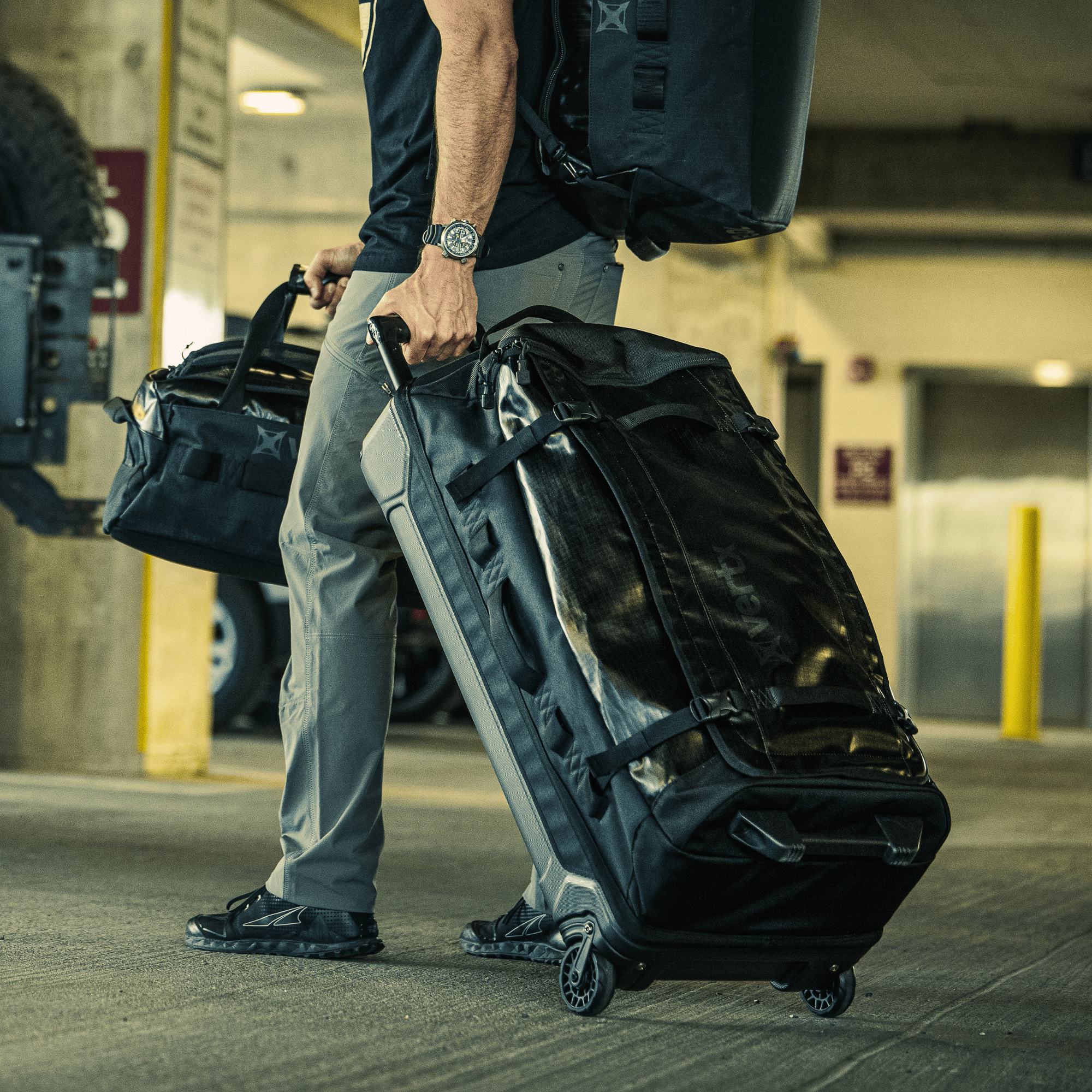 The Vertx RLT 100L Duffel Bag Roller is a black tactical duffel featuring sleek wheels and a glossy exterior, crafted for rugged durability. It includes a sturdy handle, multiple straps, side pockets, and a reinforced base to ensure enhanced mobility and longevity.