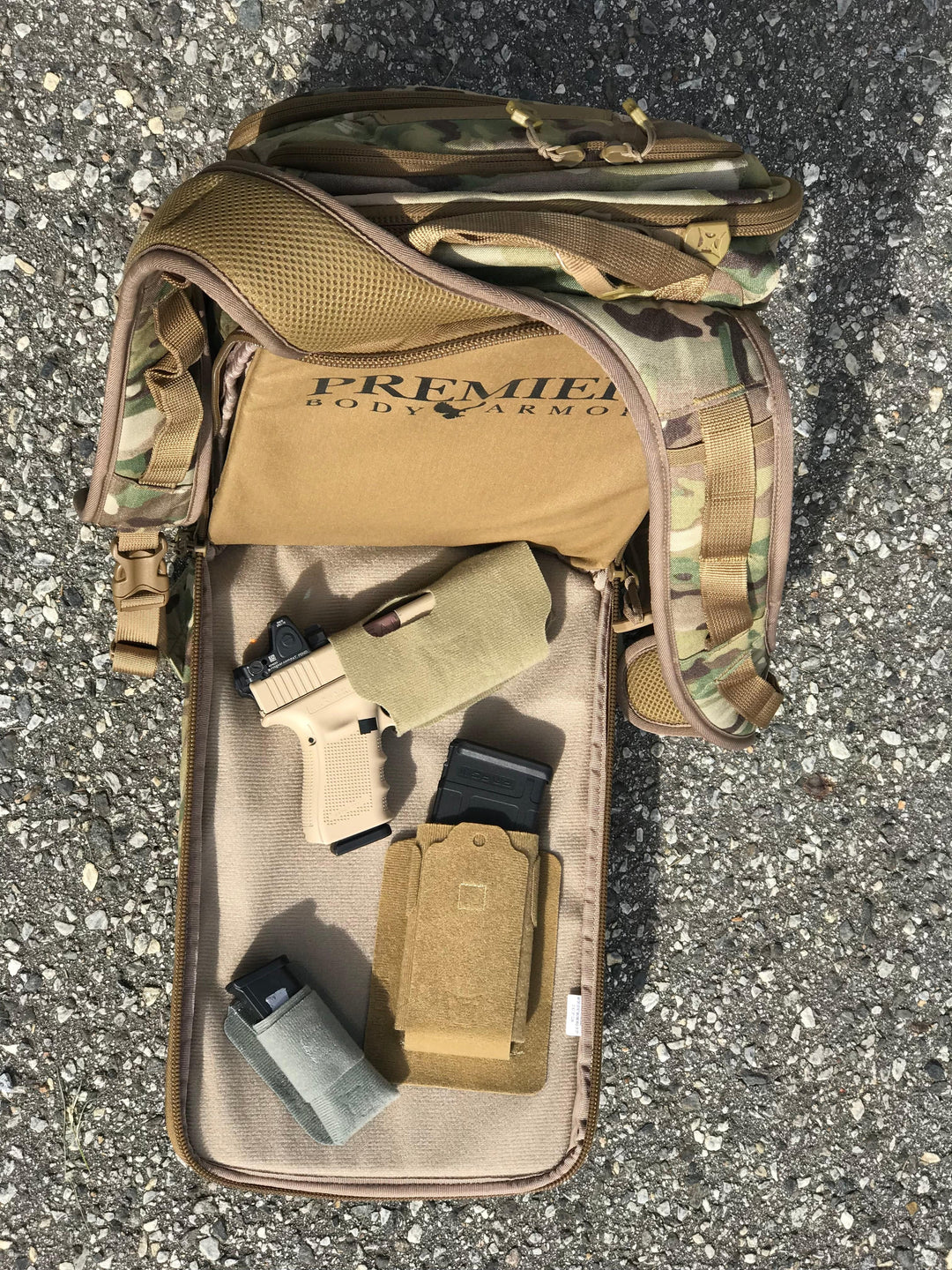 A tan and camouflage backpack with "Premier Body Armor" printed on it is open, revealing a beige sub-compact handgun in a Vertx Tactigami - MPH SUB multi-purpose holster by Vertx, alongside two ammunition magazines. The items are spread out on a gravel surface.