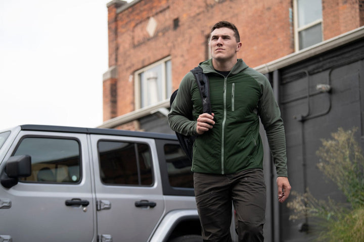 Lifestyle picture of Vertx Manitou Jacket in green. Model is carrying a bulletproof backpack. 