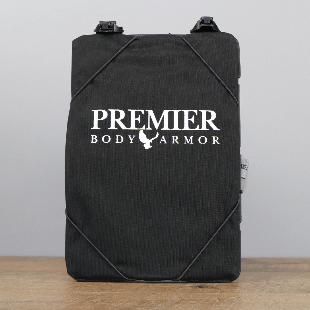 A Vertx black rectangular body armor plate with the text "Premier Body Armor" and a chicken logo printed in white. The MOLLE-compatible Vertx S.O.C.P. Panel has black straps attached and is placed upright on a wooden surface against a plain grey background.