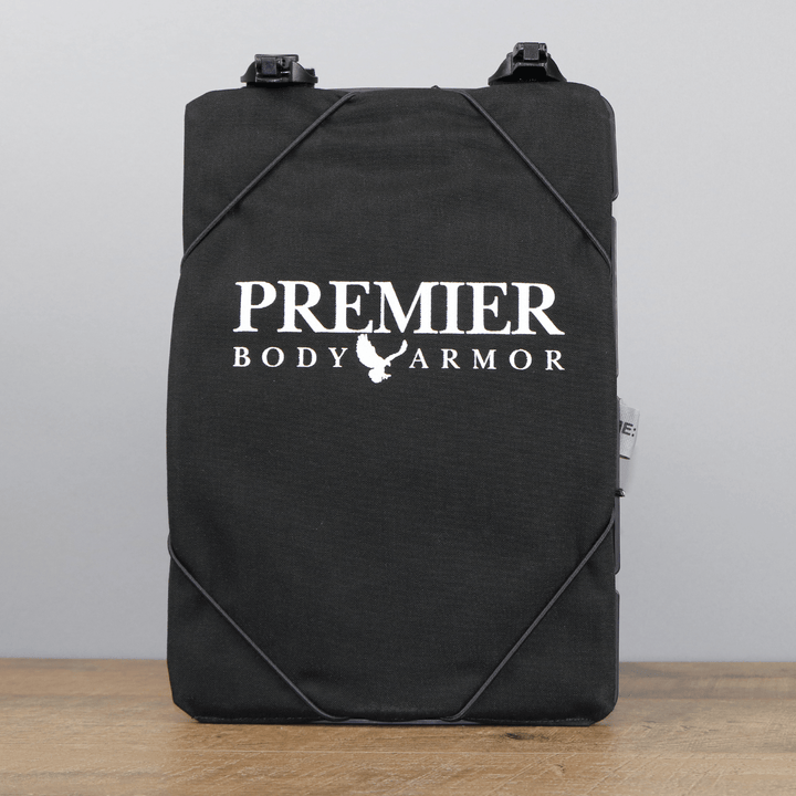 A Vertx black rectangular body armor plate with the text "Premier Body Armor" and a chicken logo printed in white. The MOLLE-compatible Vertx S.O.C.P. Panel has black straps attached and is placed upright on a wooden surface against a plain grey background.